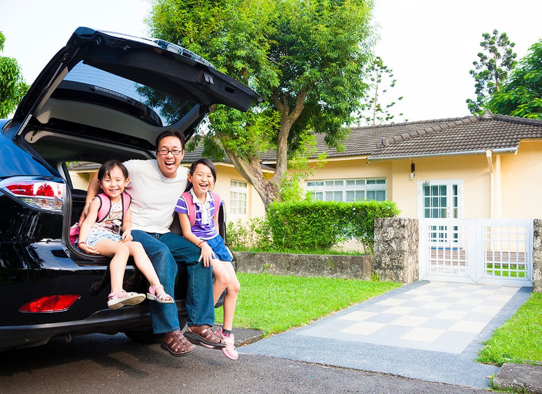 Personal Insurance - Cheerful Father Sitting in the Back Trunk of an SUV with his Two Daughters While Parked in Front of Their Single Story Home